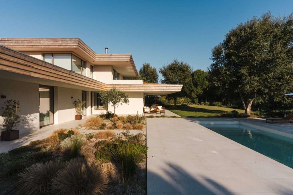 South facade of Casa da Encosta displaying extended cantilevered concrete eaves, wood trim, an outdoor pool, and local landscape gardens.