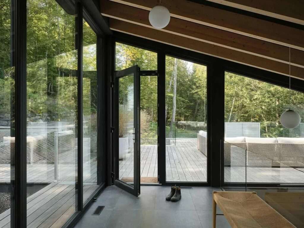 Interior dining and living area with a long light oak wooden table and chairs, a dark green sofa, and expansive floor-to-ceiling windows showing a panoramic lake view through trees.