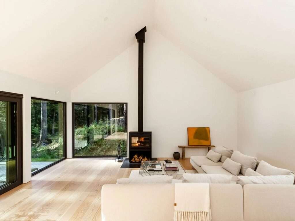 Wide-angle view of the bright, minimalist interior space under a high gabled ceiling, showing a white sectional sofa, a glass coffee table, a black fireplace chimney pipe, and expansive windows framing the surrounding forest landscape.