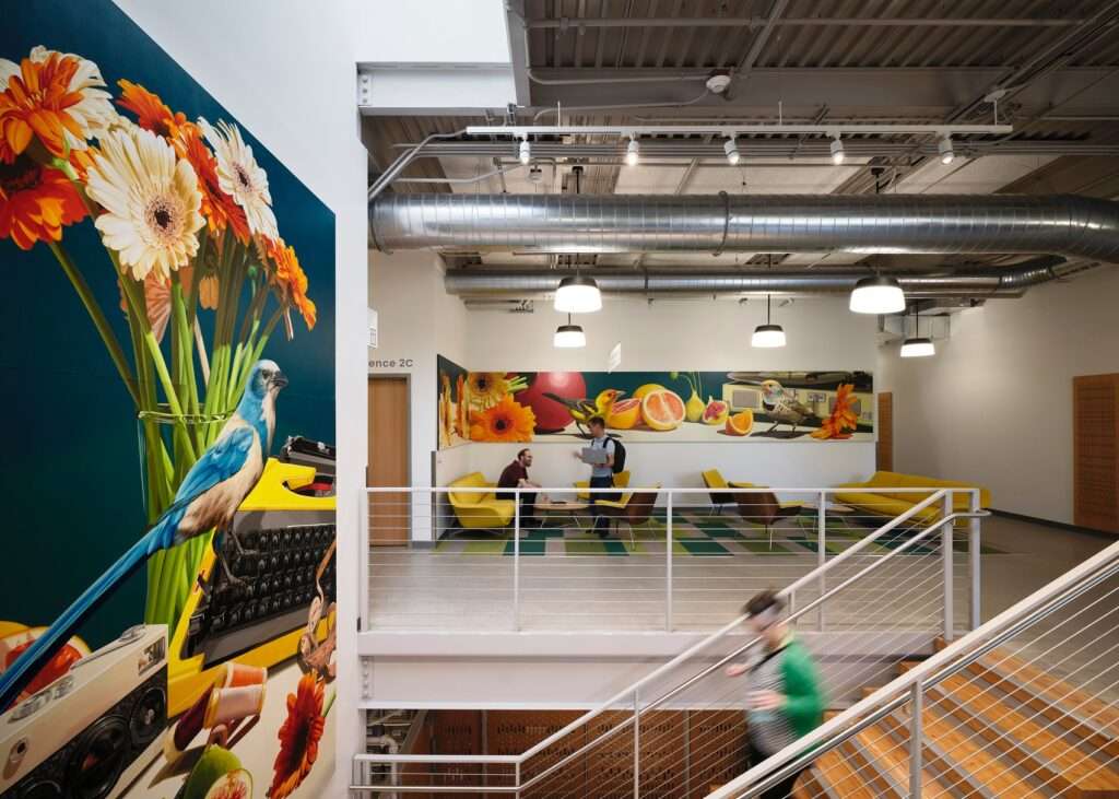Interior upper lobby of the operations center featuring a bright lounge area with yellow seating, large colorful wall murals of flowers and birds, an industrial steel staircase, and exposed ceiling ducts.