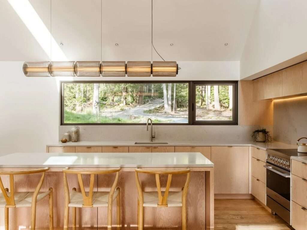Minimalist modern kitchen with custom Douglas fir wood cabinetry, a large kitchen island with wooden bar stools, white countertops, a sleek cylindrical glass pendant light fixture, and a horizontal window looking out to the forest path.