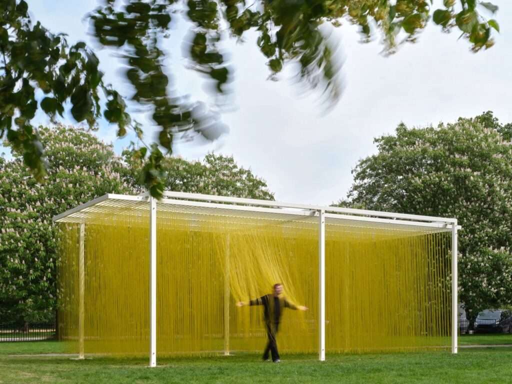 Long shot of a person moving through the center of the yellow interactive sculpture by Jesus Rafael Soto, framed by green park trees and a dynamic sky.