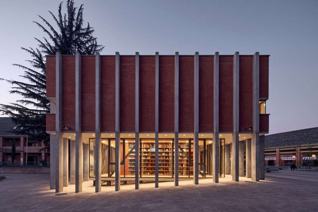 Night view of the library pavilion glowing like a central lantern, illuminating the internal bookshelves through its transparent glass ground level.
