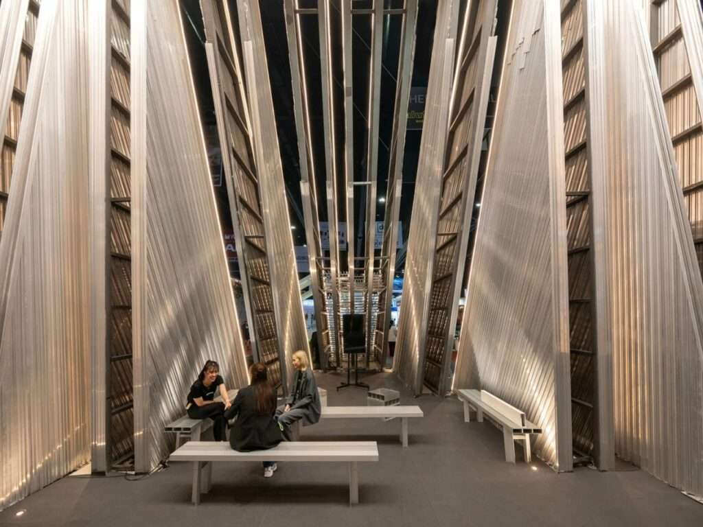 Interior of the UNFOLD Pavilion featuring linear aluminum slats forming a semi-permeable enclosure with visitors sitting on minimalist benches.