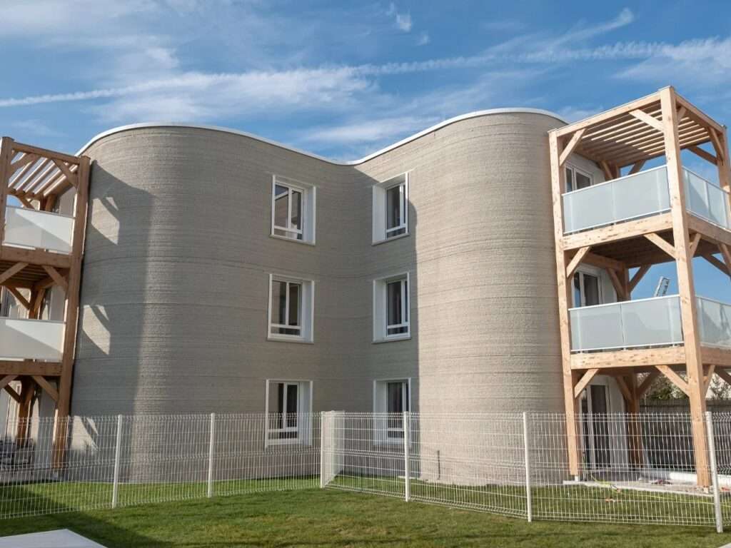 Low-angle exterior shot of the completed three-story ViliaSprint2 social housing building with fluid curved facades against a blue sky.