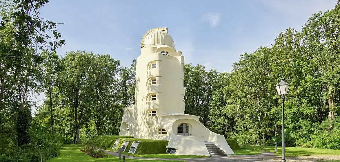 Wide panoramic landscape photograph of the iconic Einstein Tower surrounded by lush summer trees and manicured green lawns under a clear sky.