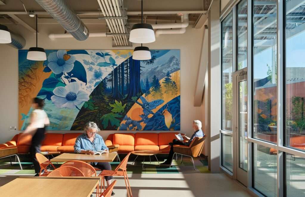 A bright staff break room filled with natural daylight coming through floor-to-ceiling windows, orange lounge seating, wood furniture, and a panoramic nature-themed mural.