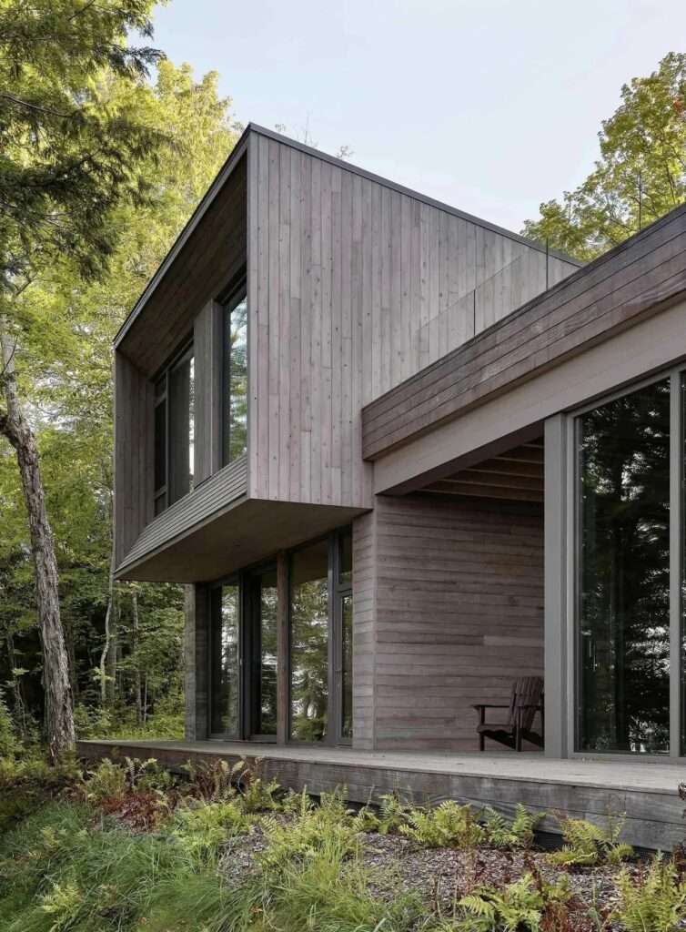 A modern minimalist kitchen featuring light oak cabinetry, light grey stone countertops, and a large window looking out onto a steep forested slope.