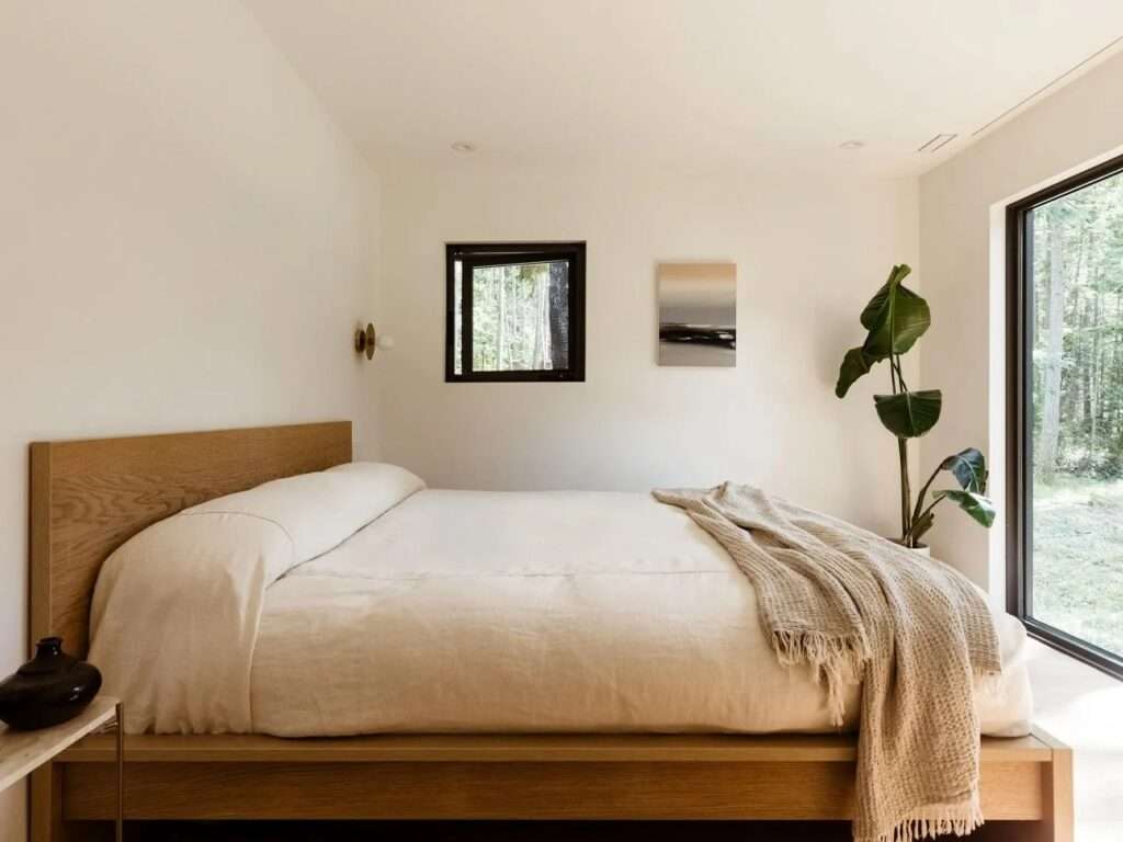 Serene minimalist bedroom featuring a wooden bed frame with crisp white linens, a large picture window overlooking the dense forest, a small square ventilation window, a potted banana plant, and soft ambient lighting.