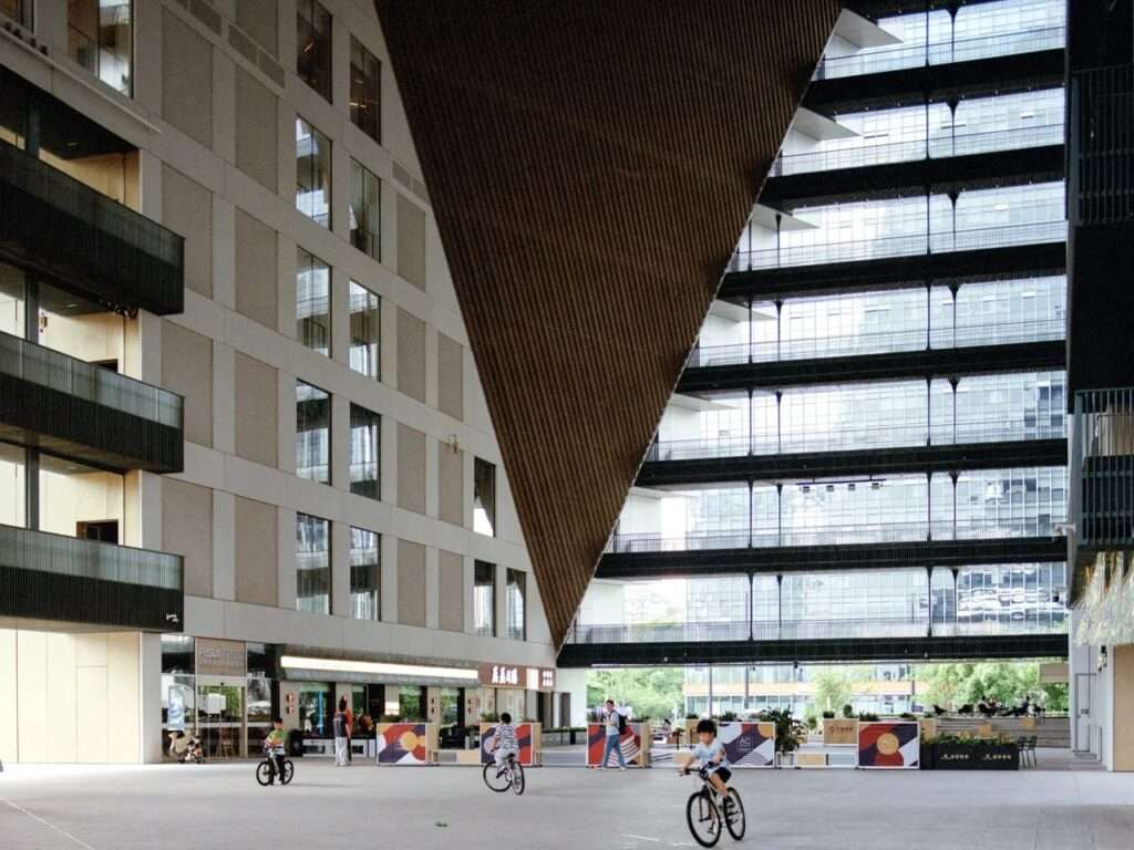 Large open-air public ground plaza under the massive diagonal cantilever of Hangzhou Prism with people riding bicycles.