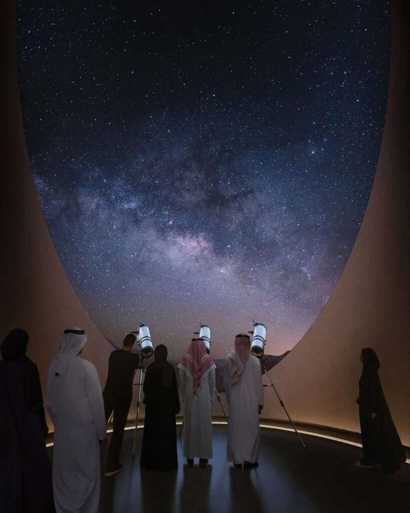 Interior of AlUla Manara observatory showing visitors in traditional attire using telescopes to view the starry night sky through a massive oval ceiling opening.