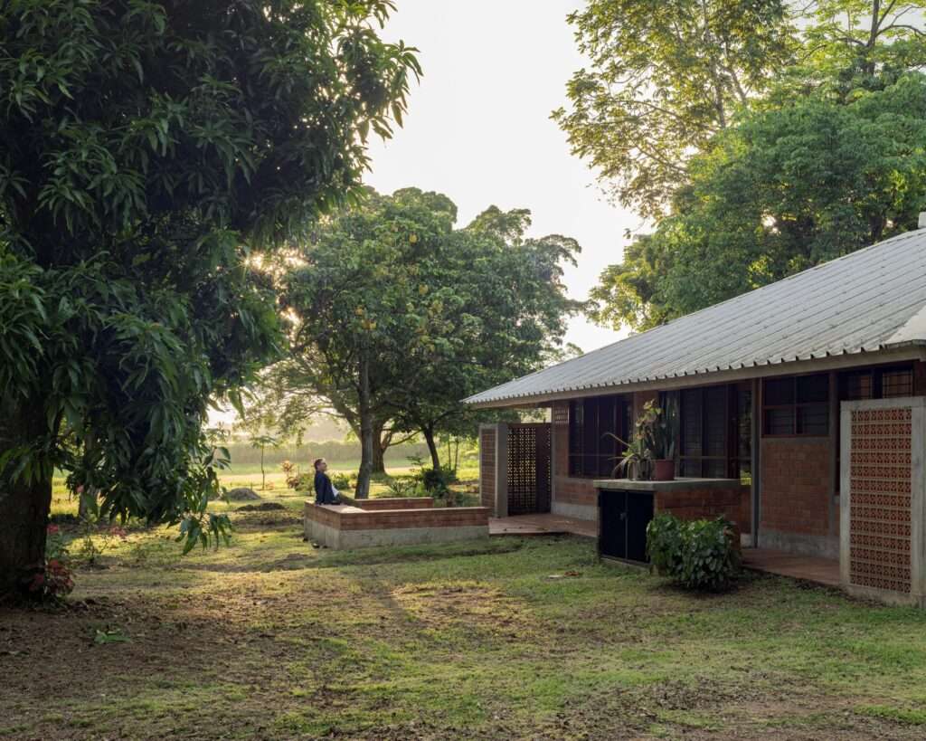 South facade of Villa E during golden hour, revealing a brick-clad home with wide roof eaves and a resident sitting on a low brick retaining wall outside.