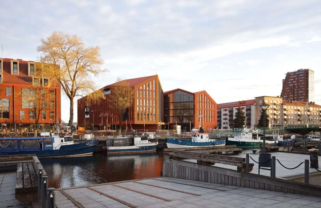 Distant waterfront view of the contemporary terracotta HANZA building sitting along the canal in Klaipeda, surrounded by historic brick buildings and docked fishing boats.
