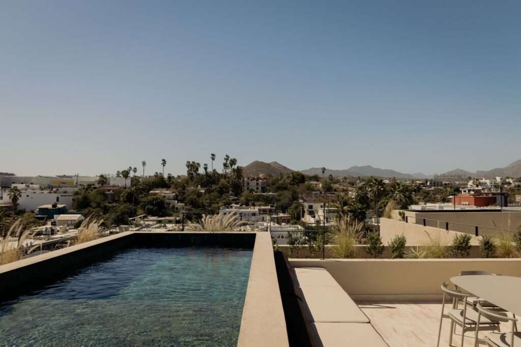 Rooftop swimming pool terrace at Estero Residences overlooking the historic cityscape of San José del Cabo, surrounded by mountains under a clear sky.