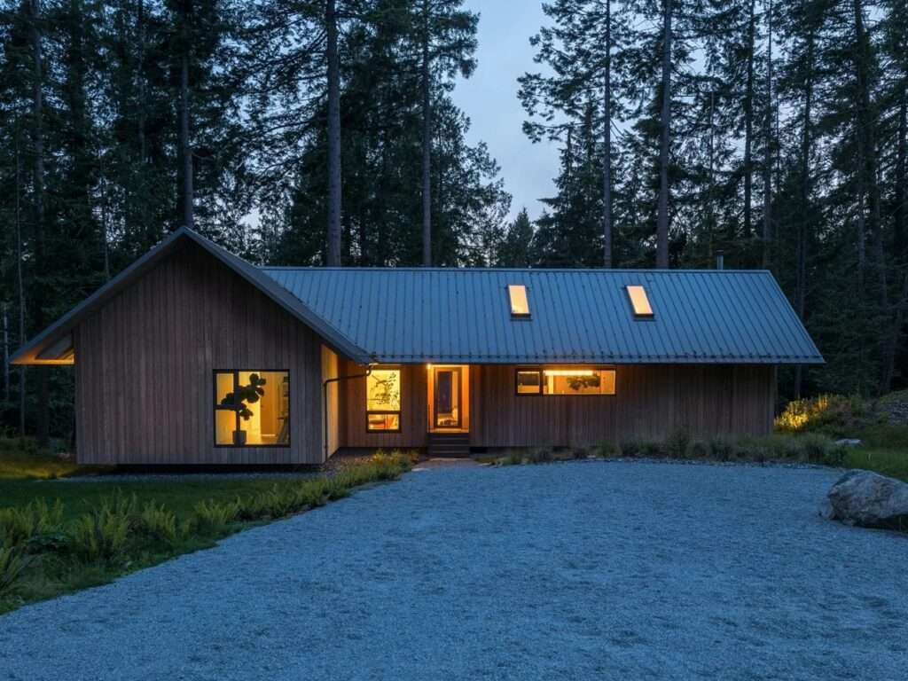 Dusk view of the Floating House exterior with warm interior lights glowing through the windows, casting a soft light over a gravel driveway and the surrounding dark evergreen forest of Bowen Island.