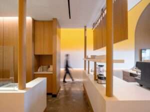A perspective view through a series of wooden columns that frame the long white service counter at Fuwa Fuwa. A blurred person in motion walks past the counter, set against the warm yellow curved ceiling.
