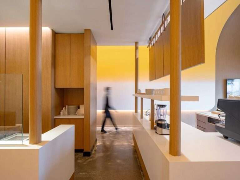 A perspective view through a series of wooden columns that frame the long white service counter at Fuwa Fuwa. A blurred person in motion walks past the counter, set against the warm yellow curved ceiling.