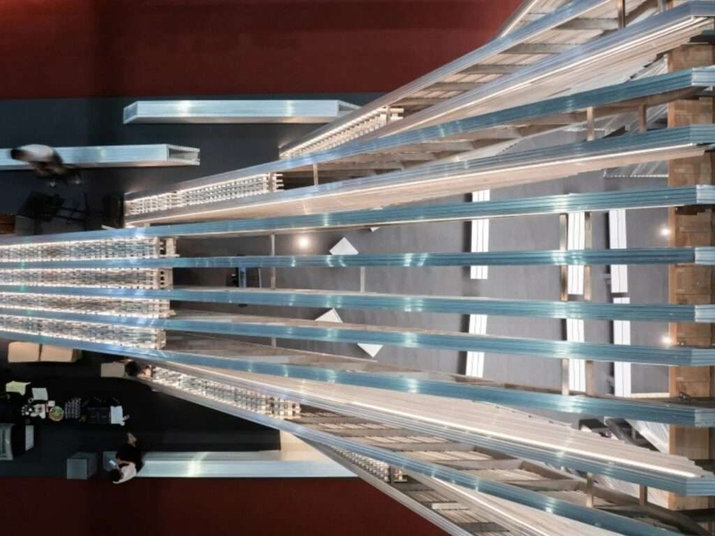 Abstract top-down view looking down the length of the UNFOLD Pavilion's linear aluminum beam system over the gray exhibition floor.