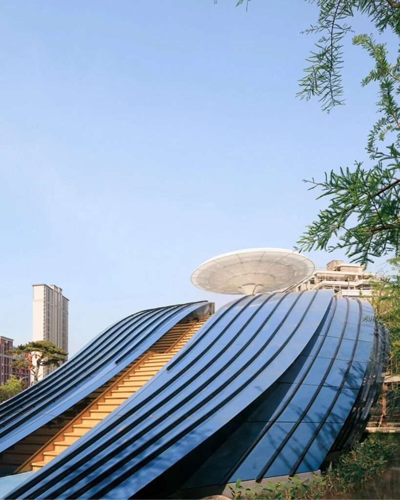 Aerial top-down view of the Whale Gate pavilion featuring a blue curved aluminum shell, a stone path, and an upper viewing deck with a white circular umbrella canopy.