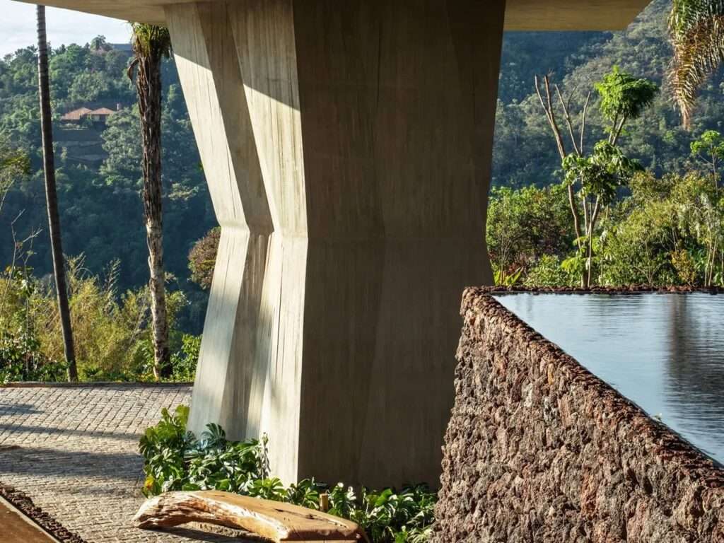 Monolithic angular concrete pillar next to a rustic volcanic stone retaining wall and an infinity edge pool.