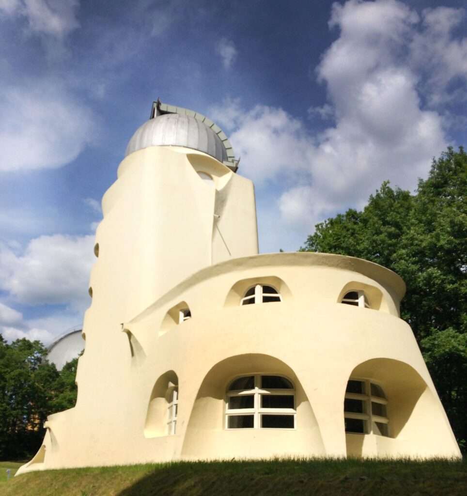 Low-angle view looking up at the curved rear tiers of the Einstein Tower with its distinct arched windows against a vibrant blue sky with scattered clouds.