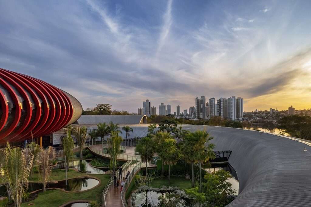 Panoramic dusk view of the Pantanal Biopark, highlighting the oval zinc-clad building surrounded by landscaped ponds and city skyline in the background.