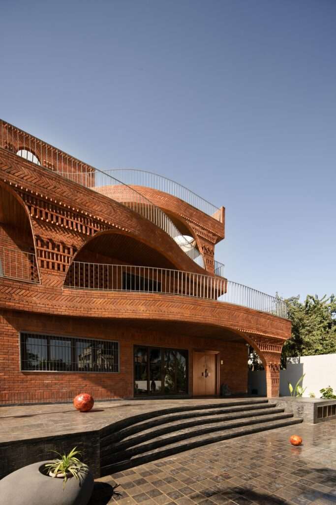 Exterior view of the building's side facade showing brick jaali screens, multi-layered terraces with potted plants, and a partial view of a palm tree.