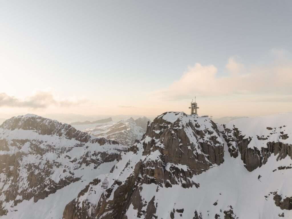 Distant wide view of the isolated Titlis tower standing atop a jagged, snow-dusted alpine ridge under a hazy sky.