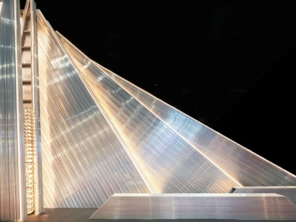 Clean profile view focusing on the layered, sloped aluminum segments of the UNFOLD Pavilion against a stark dark background.