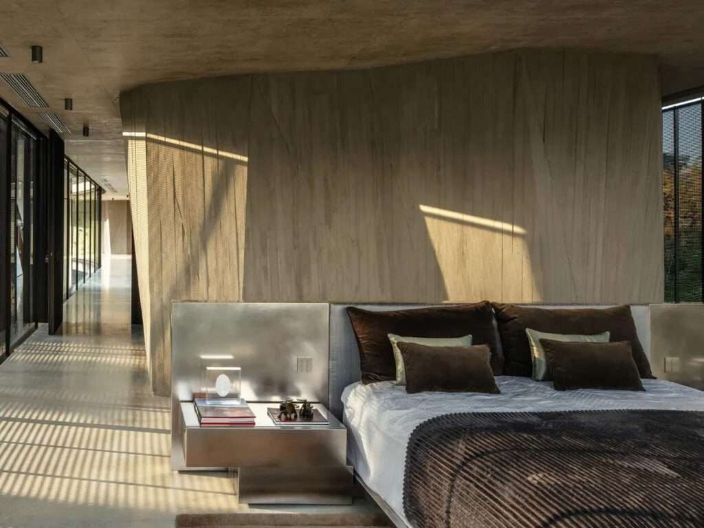 Minimalist luxury bedroom inside Xingu House with raw concrete walls, a metallic headboard, and warm sunlight casting shadows.