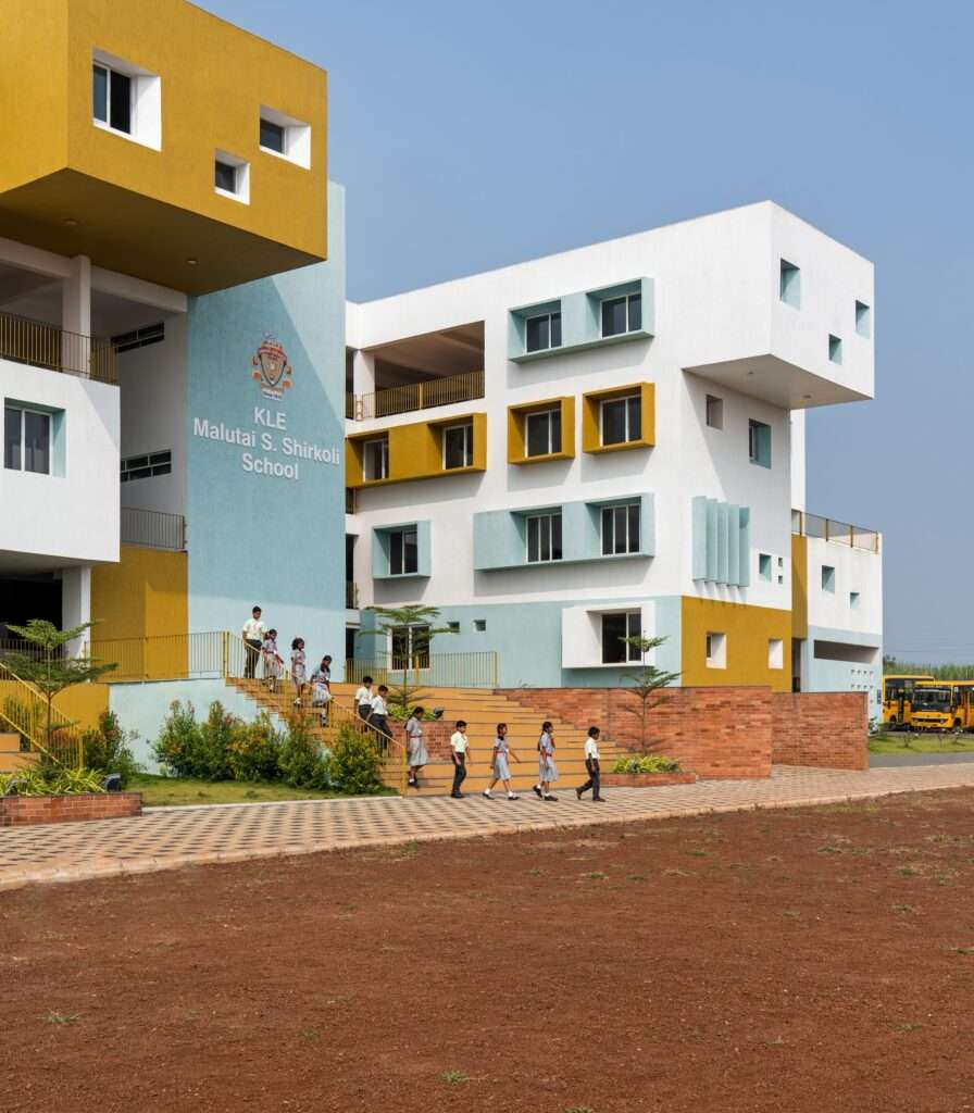 Corner view of a contemporary school building showing white, cyan, and mustard yellow block modules with students walking down outdoor steps flanked by brick walls.