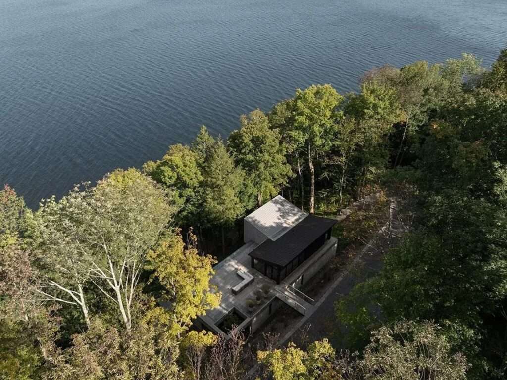 A side perspective of a modern sloping-roof house built on a concrete foundation wall with a wooden bridge crossing a natural, grassy ravine flanked by tall trees.