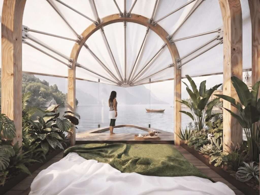 Interior view of the Lucia floating home showing a bedroom space with a low bed, lush green plants on the sides, glass walls, and a woman looking out at Lake Como through an arched wooden frame.