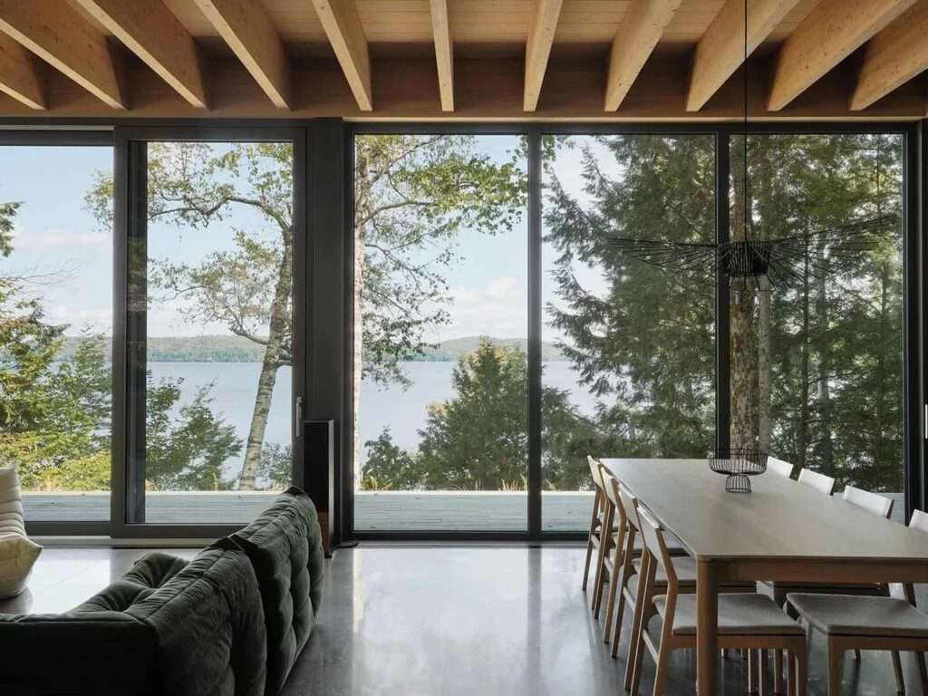 Interior view looking out through black-framed glass walls to an exterior wooden deck with an open door, featuring exposed ceiling timber beams and minimalist furniture.