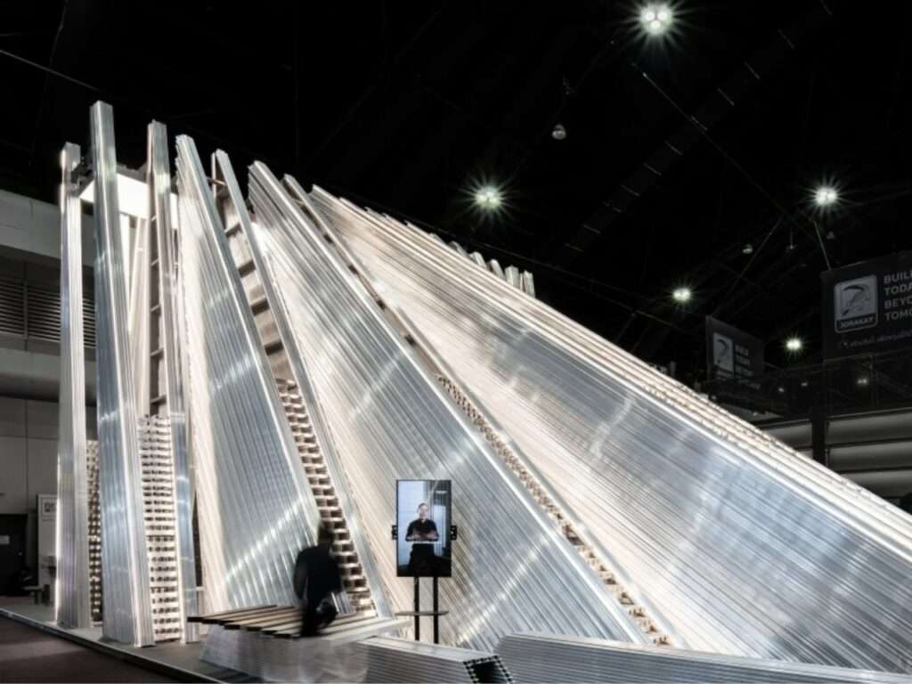 Front perspective of the UNFOLD pavilion featuring an interactive digital display screen standing beside the layered metallic structure.