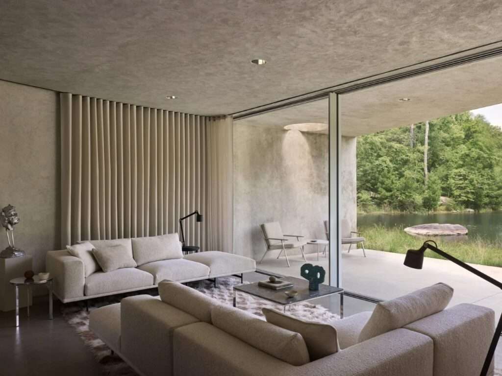 Luxury minimalist living room with floor-to-ceiling glass windows overlooking a pond and forest.