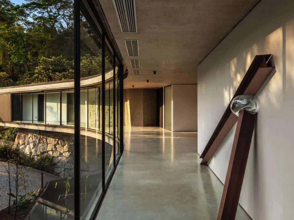 Long minimalist glass corridor inside Xingu House showing a curved glass facade overlooking stone ruins and lush trees.