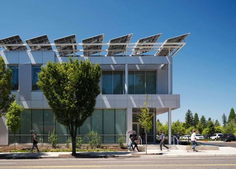 Close-up of the two-story building facade showing metallic paneling, clean window lines, a street-facing sidewalk with pedestrians, and angled rooftop solar panels casting crisp shadows.