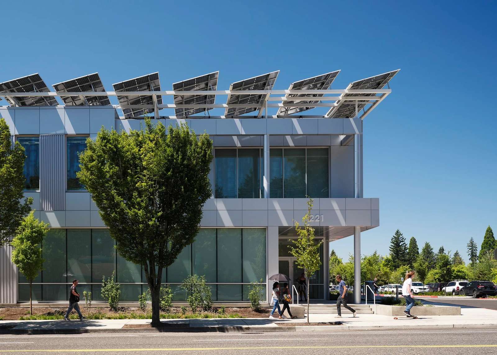 Close-up of the two-story building facade showing metallic paneling, clean window lines, a street-facing sidewalk with pedestrians, and angled rooftop solar panels casting crisp shadows.