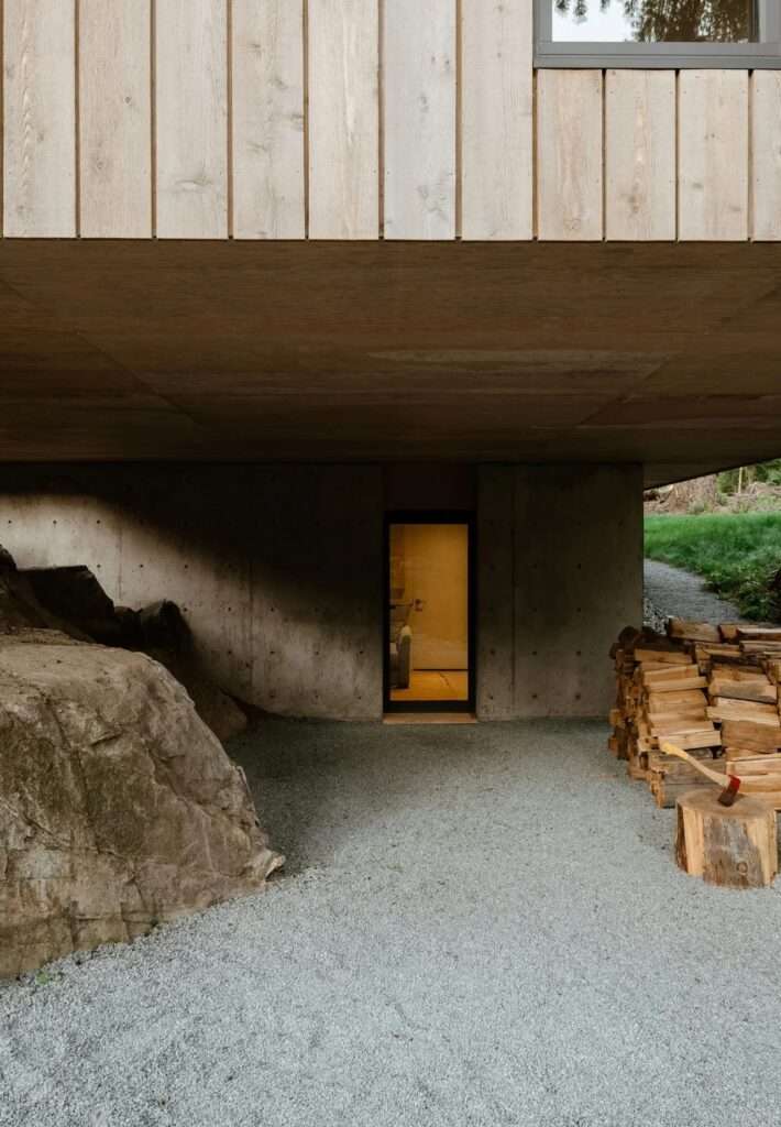 Underneath the cantilever of the Floating House, showing a gravel patio area adjacent to a massive natural boulder, a stack of firewood with an axe, a concrete foundation wall, and a glowing glass entry door.