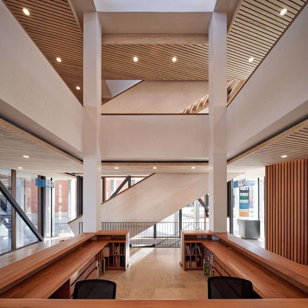Symmetrical view from a central reception desk looking up at a double-height atrium with structural concrete columns, wooden staircases, and a skylight.