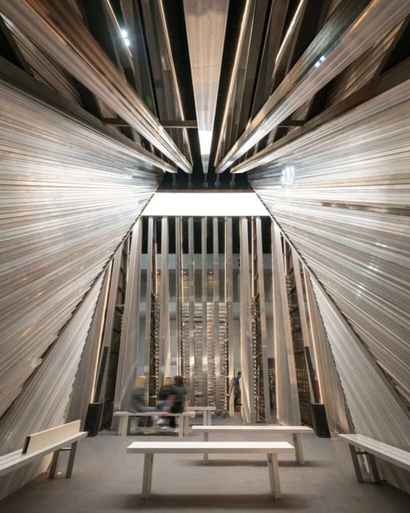 Looking up inside the UNFOLD pavilion toward a dramatic converging ceiling canopy made of repetitive aluminum slats and a central skylight feature.