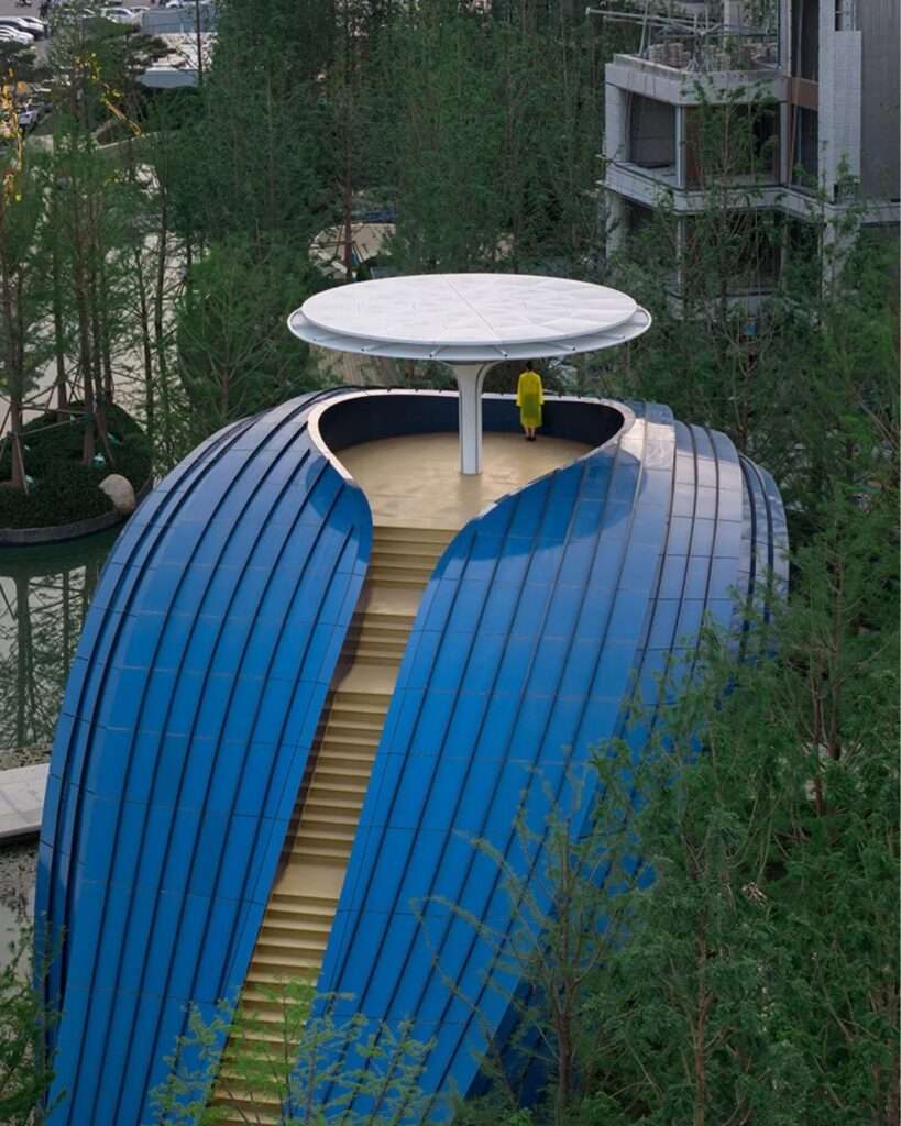 Close-up detail of the golden metal steps positioned between the overlapping blue double-curved aluminum cladding panels of the pavilion.