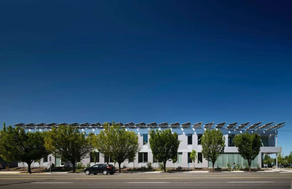 Full horizontal street view of the long metallic and glass facade of the Multnomah County Library Operations Center with a continuous row of rooftop solar arrays, green trees, and a passing car.