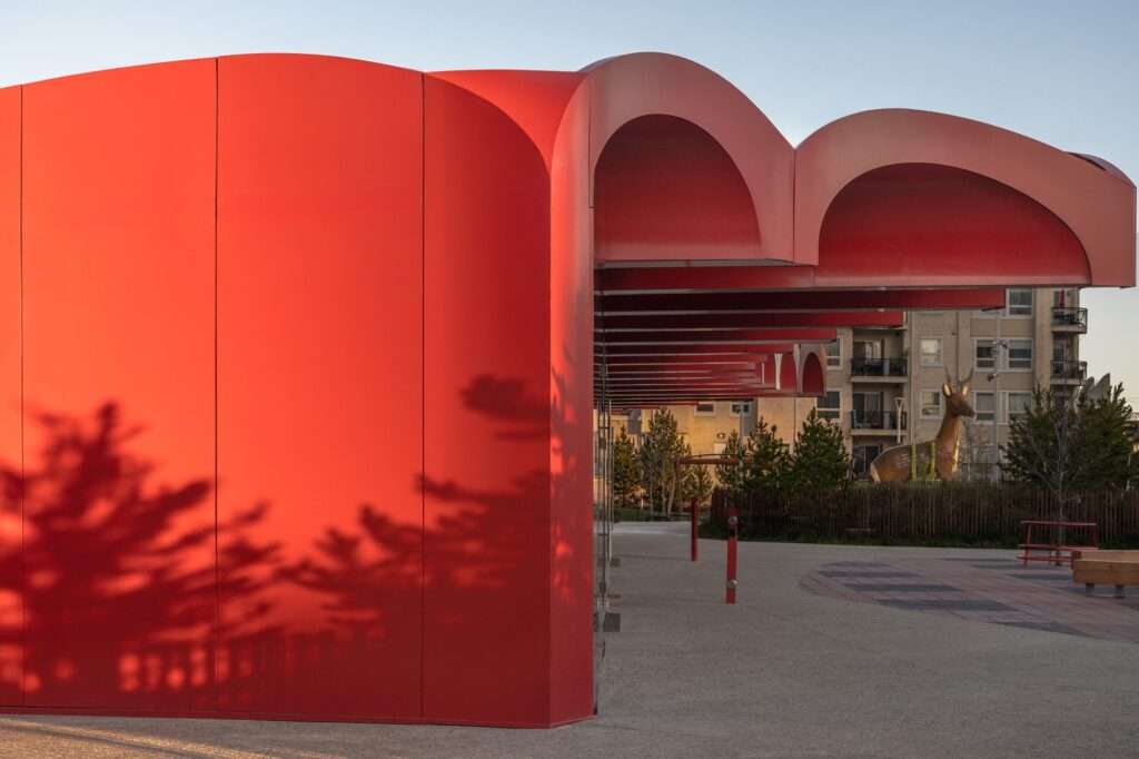 Side view of the O-day'min Park Pavilion canopy in Edmonton, showcasing the deep red facade and the seamless integration into the surrounding park landscaping.