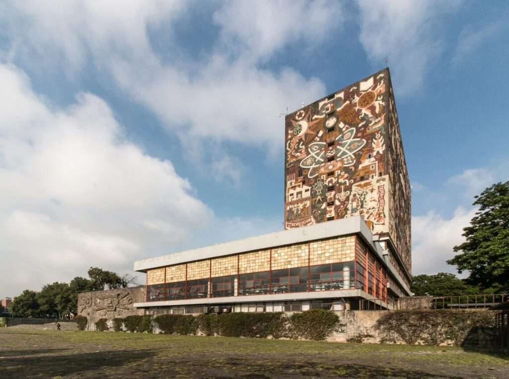 Wide angle view of the open stone plaza at Ciudad Universitaria showing the UNAM Central Library in the distant background, a volcanic rock sculpture in the foreground, and the high-rise Rectorate Tower on the left.