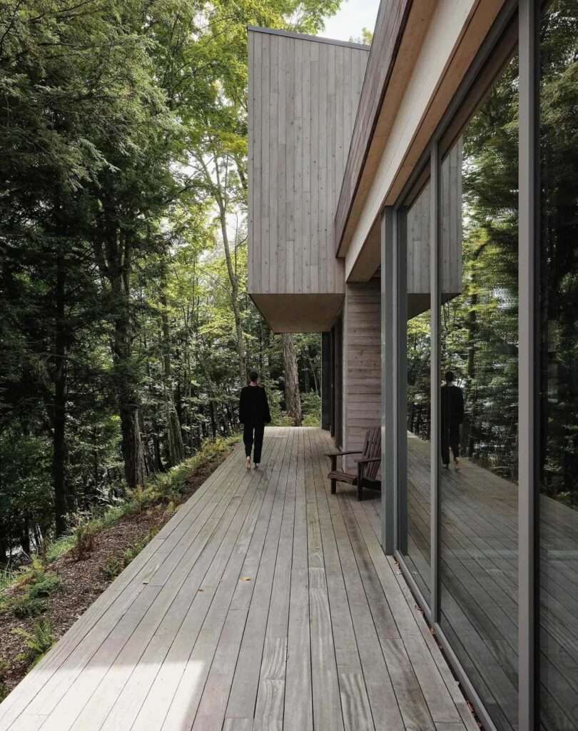 Wide exterior wooden deck of a modern house overlooking a forest, featuring an outdoor sofa, glass railings, and a black-framed angled roofline.