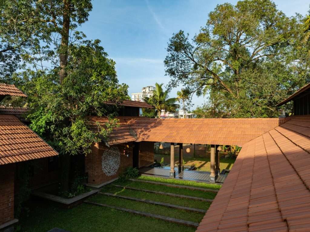 High-angle rooftop view looking down over interlocking clay tiled roofs into a stepped green courtyard framed by mature mango trees.
