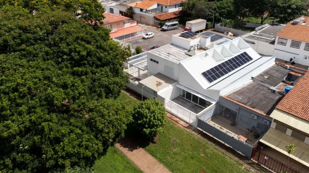 High angle aerial drone view of a narrow white modern house with solar panels on its sloped roof, situated within a dense urban neighborhood in Brasilia next to green spaces.
