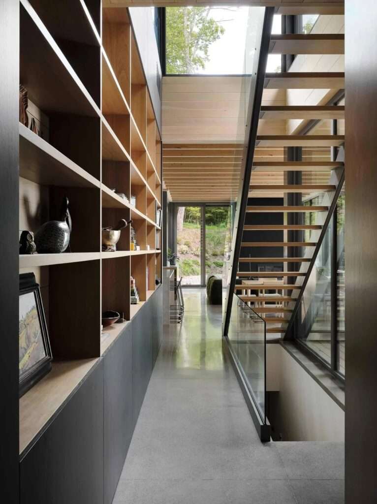 Interior hallway featuring a tall floor-to-ceiling wooden bookshelf on the left and an open-riser wooden staircase with glass railings on the right, leading down to a polished concrete floor.